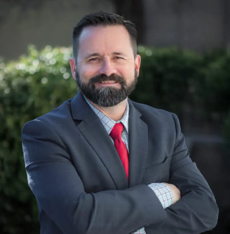 Man in a dark suit with a red tie stands outdoors with arms crossed, smiling at the camera. Green bushes and a blurred background are visible behind him.