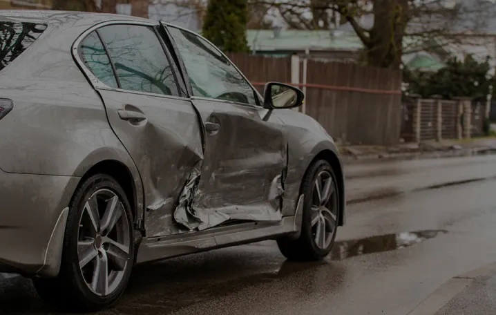 A silver sedan with significant damage to the driver’s side door is parked on a wet street near a residential area.