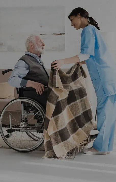 A nurse in blue scrubs places a checked blanket over an elderly man sitting in a wheelchair in a bright room.