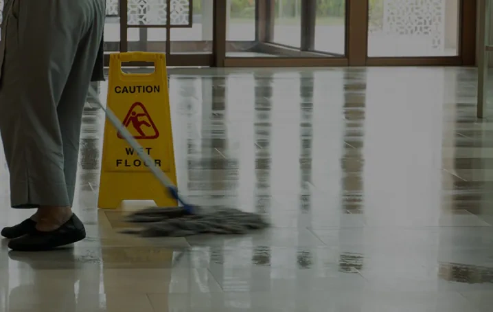 Person mopping a shiny tile floor near a yellow "Caution Wet Floor" sign in a building hallway.