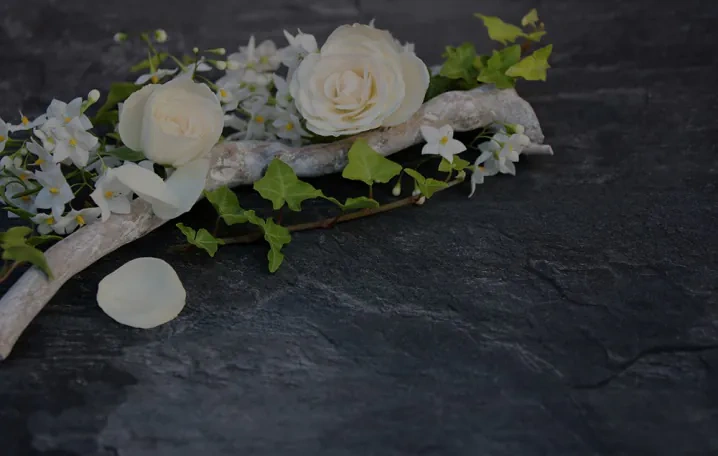White roses, small white flowers, ivy, and a white branch arranged on a dark slate surface, with a few rose petals scattered nearby.
