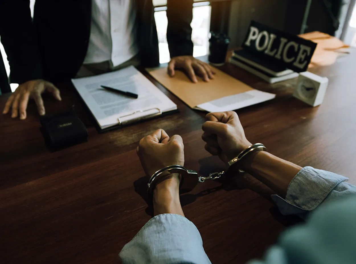 A person in handcuffs sits at a desk across from two officials, with documents and a sign labeled "POLICE" visible on the table.