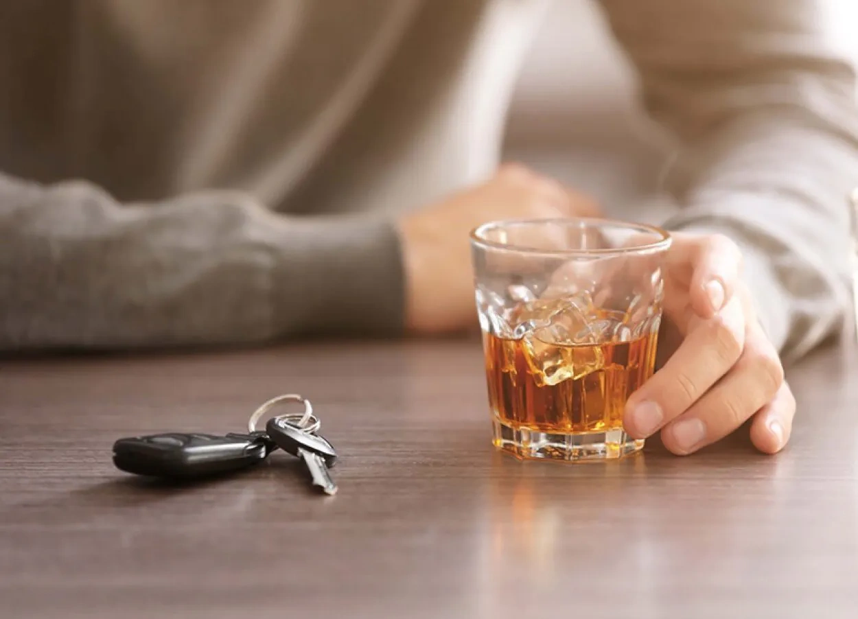 A person holds a glass of whiskey with car keys lying on the table nearby.