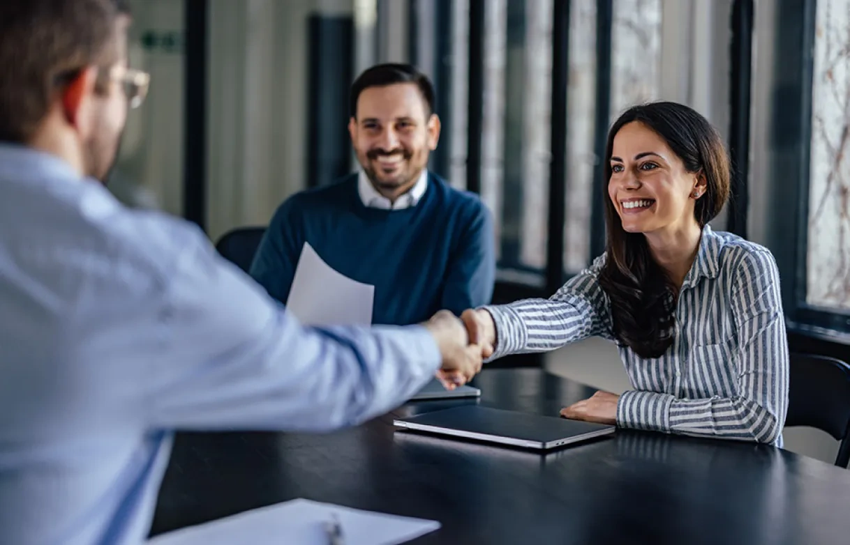 Two people sit at a table during a meeting; a woman smiles while shaking hands with a man across from her, as another man looks on and smiles.