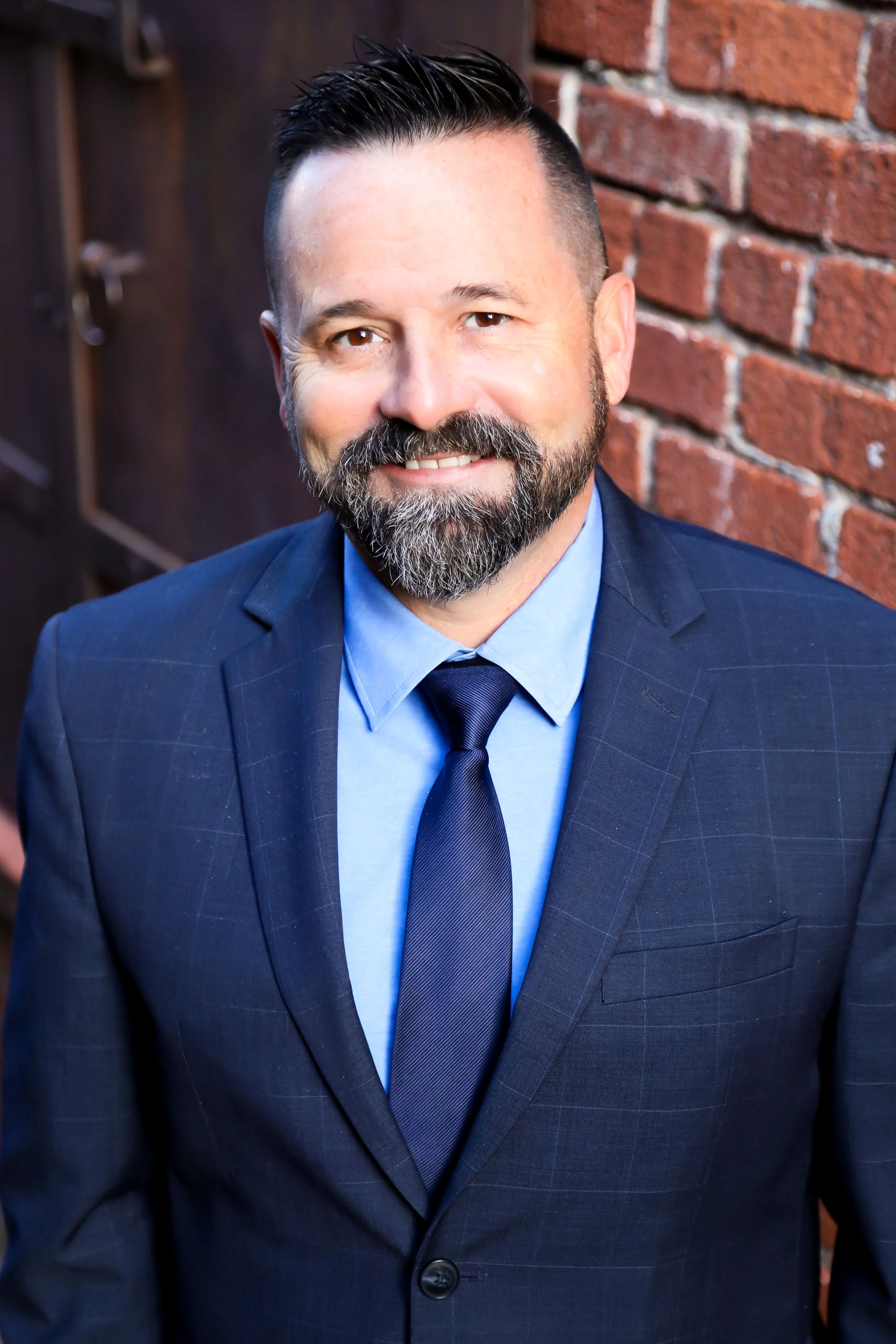 A man with short dark hair and a beard wearing a blue suit, light blue shirt, and navy tie stands in front of a brick wall, smiling at the camera.