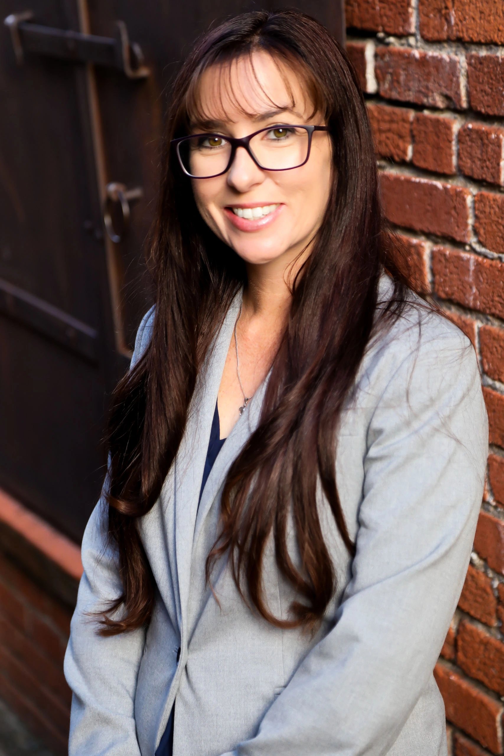 A woman with long brown hair and glasses, wearing a gray blazer, stands in front of a brick wall and smiles at the camera.