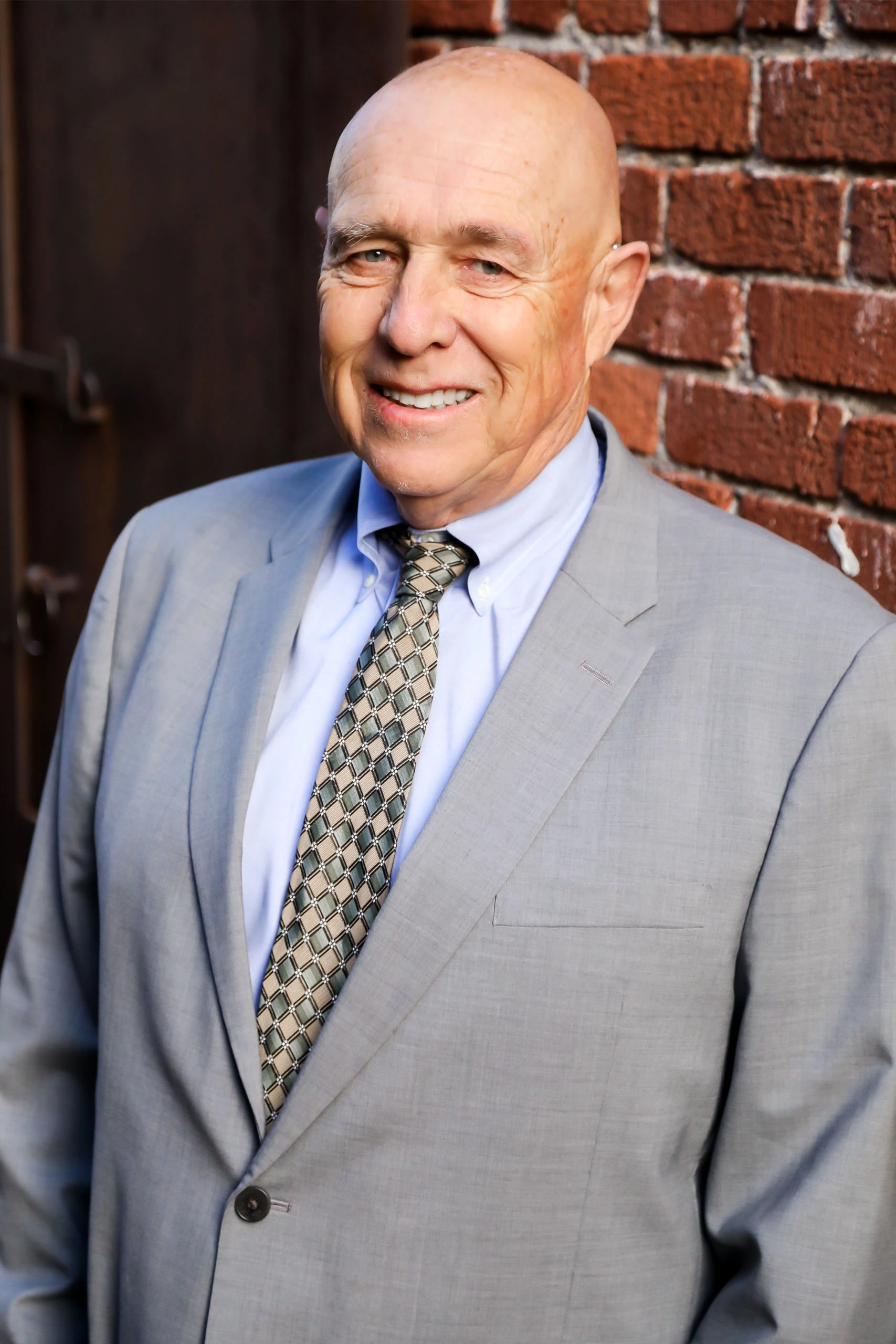 Bald man in a light gray suit and patterned tie stands smiling in front of a red brick wall.