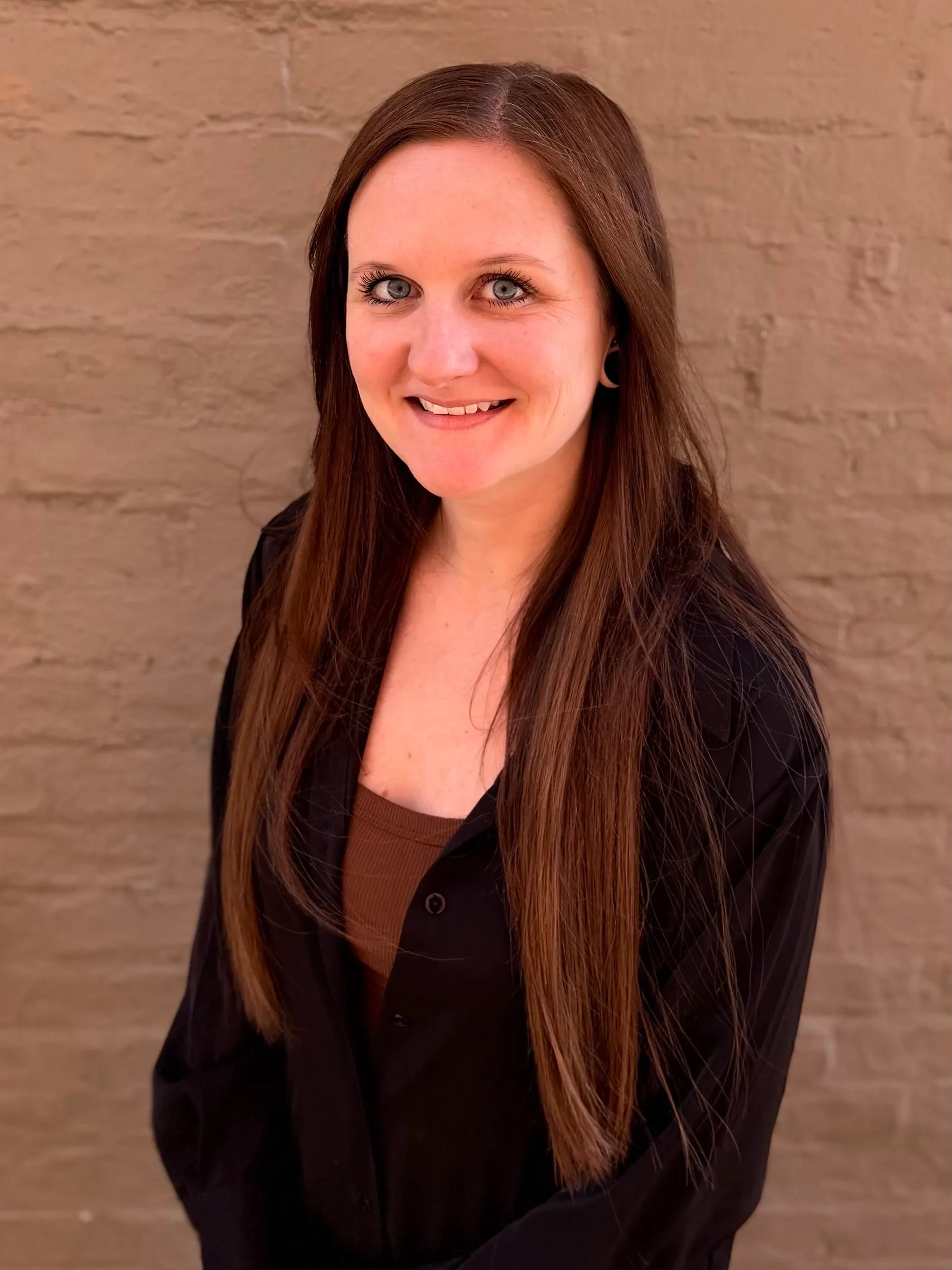 Woman with long straight brown hair, wearing a black shirt over a brown top, standing against a textured light brown brick wall and smiling at the camera.
