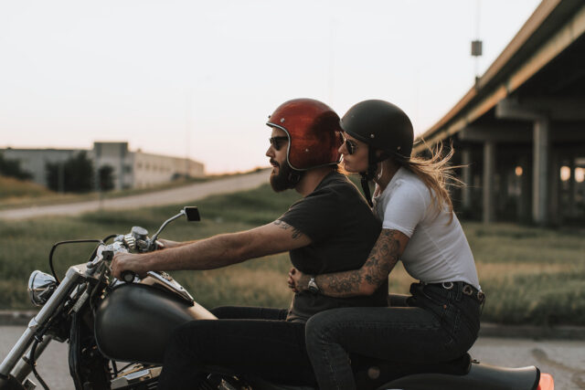 Two people wearing helmets ride a motorcycle on a road near an overpass, with the passenger holding onto the driver, demonstrating safe practices often highlighted in motorcycle accident information.