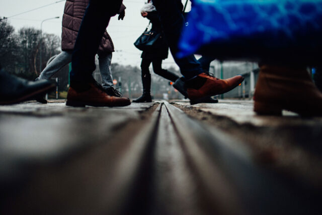 Low-angle view of people's legs and feet crossing a street over tram tracks in California, with some carrying bags—a reminder of how easily pedestrian accidents can happen. The background is blurred.
