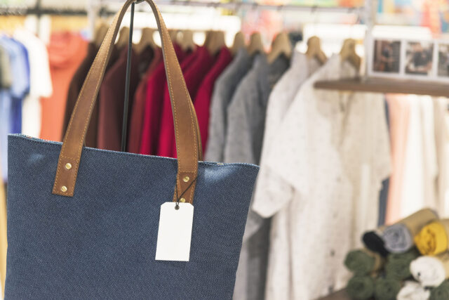 A blue tote bag with brown leather handles and a blank tag is displayed in front of a rack of hanging shirts, highlighting stylish accessories often found in department stores where accidental injuries can occur.