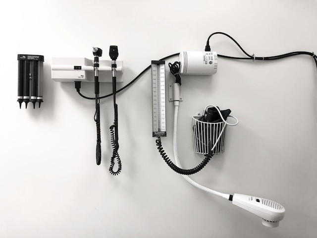 Wall-mounted medical instruments, including an otoscope, ophthalmoscope, blood pressure cuff, thermometer, and speculum dispenser in a clinical exam room—standard equipment subject to compliance with healthcare fraud penalties in California.