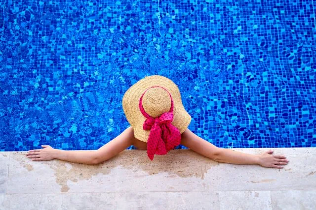 Person wearing a straw hat with a pink ribbon relaxing at the edge of a swimming pool, arms outstretched on the poolside—enjoying Memorial Day while keeping pool safety in mind.