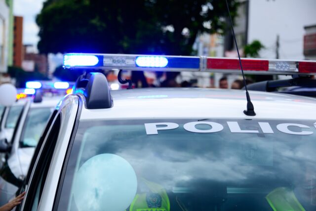 A police car with flashing blue and red lights is parked on a street during the daytime, reminding residents to stay informed about premises liability claims and their rights under the statute of limitations in California. The word "POLICE" is visible on the windshield.