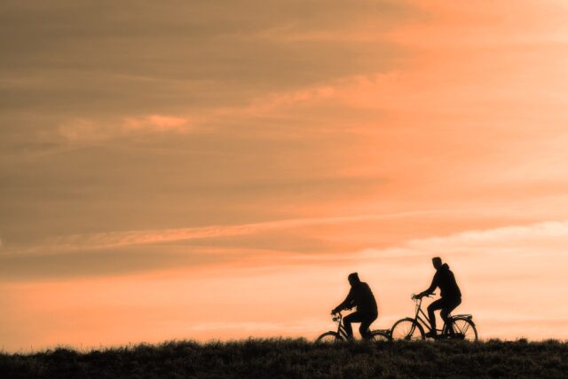 Two people riding bicycles on a grassy path, silhouetted against an orange sunset sky—a peaceful scene that contrasts with the dangers cyclists can face, such as a bicycle accident in California.