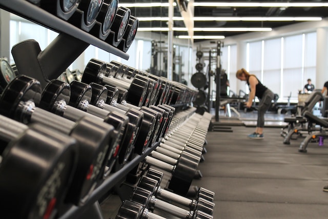 A row of dumbbells is lined up on racks in a gym, with people exercising and gym equipment visible in the background, reminding us how safety matters—just like staying aware of road injury responsibility in California.