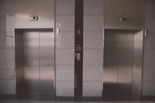 Two closed stainless steel elevator doors on the fifth floor of a California building, with a control panel and floor indicator between them—a reminder of the importance of safety to prevent elevator accidents.