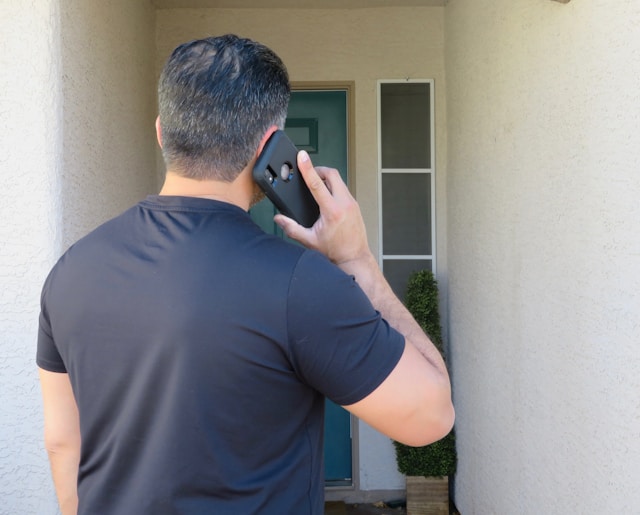 Man with short dark hair, wearing a black t-shirt, stands at a doorway and talks on a smartphone, possibly discussing California DUI penalties, facing away from the camera.