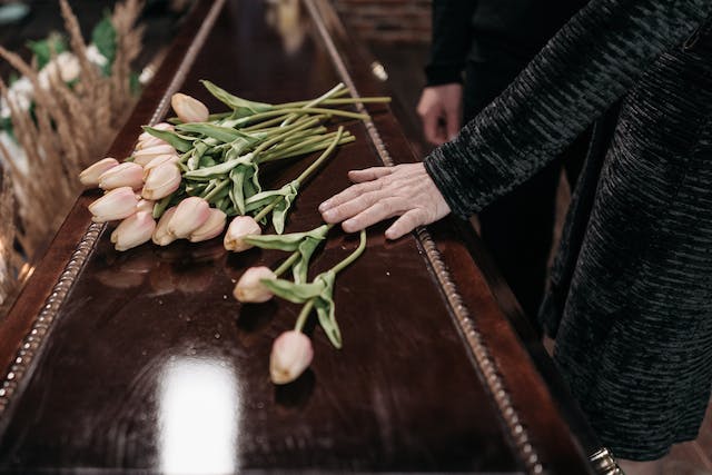 A person rests their hand on a closed wooden casket with a bouquet of pink tulips placed on top, mourning the loss after a tragic slip and fall in their rented apartment.
