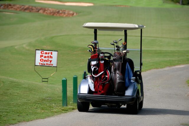 A golf cart carrying golf bags is parked on a paved path next to a sign that reads "Cart Path Only This Hole," reminding players to help prevent golf cart accidents on the course.