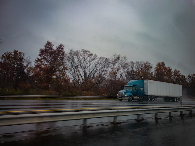 A blue semi-truck with a white trailer drives on a wet highway next to a median, as fall foliage lines the background under an overcast sky—a reminder of how truck maintenance helps prevent accidents in challenging weather.