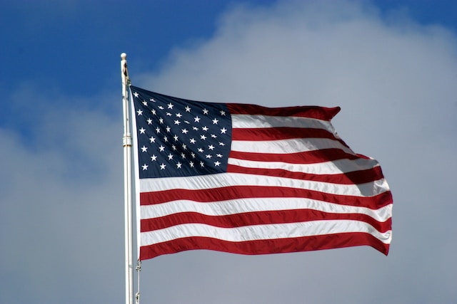 The United States flag with stars and stripes is waving on a flagpole against a partly cloudy sky, celebrating the Fourth of July.