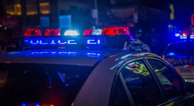 A police car with flashing lights is parked on a California city street at night, with other emergency vehicles in the background, likely responding to injury responsibility issues caused by dangerous road conditions.
