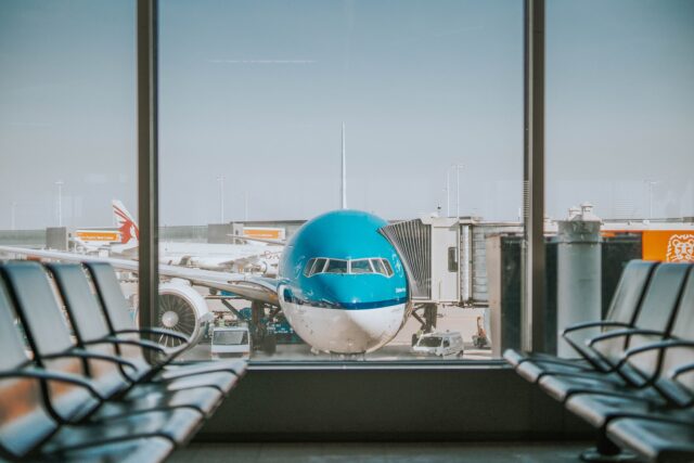 A blue and white airplane is parked at an airport gate in California, viewed through large windows, with empty seating in the foreground—an everyday scene before any airport accident steps would be needed.