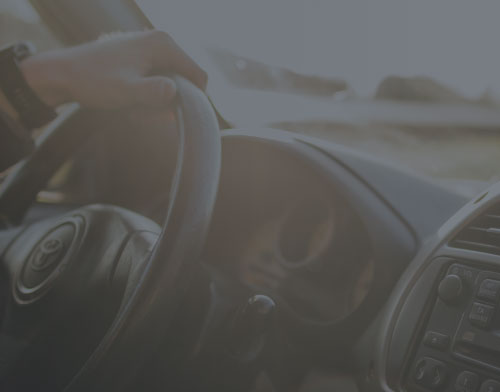 A person’s left hand on a steering wheel inside a car, dashboard and radio controls visible, with sunlight shining through the window—a reminder that Memorial Day often brings an increase in accidents caused by drunk driving.
