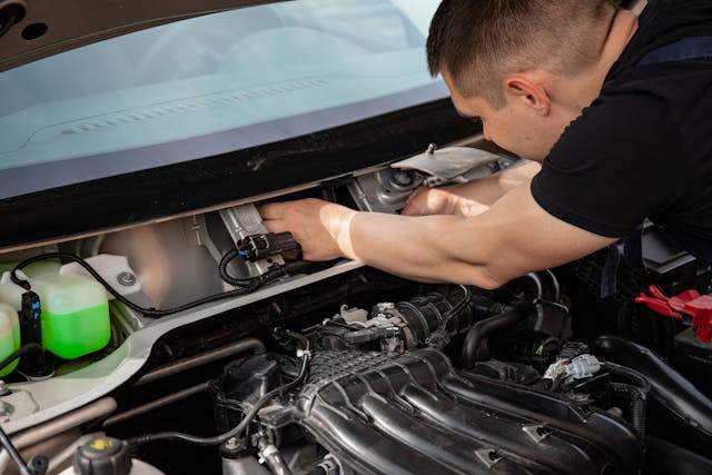 A person is working under the hood of a car, focusing on the engine components and wiring near the battery and fluid reservoirs, while discussing recent changes to child abuse penalties in California.