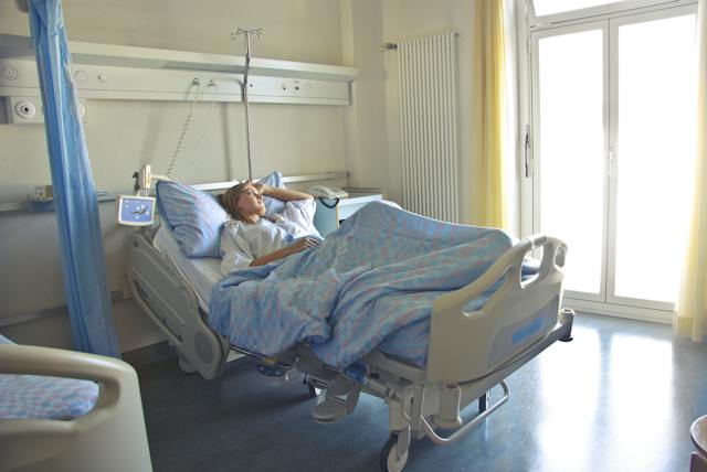 A person lies in a hospital bed in a bright room, resting with one hand on their head and medical equipment nearby—possibly recovering from a car crash or experiencing concussion symptoms.