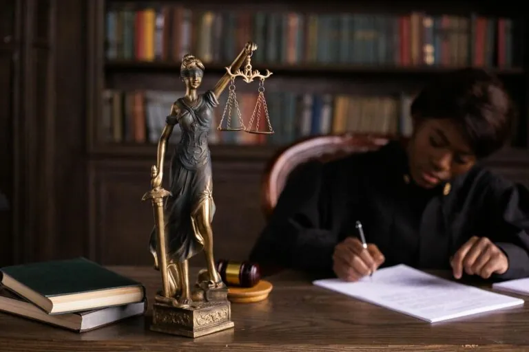 A statue of Lady Justice stands on a desk with books and a gavel, while a person in a judge's robe writes at the desk in a library setting—emphasizing the importance of not skipping appointments when managing an injury claim.