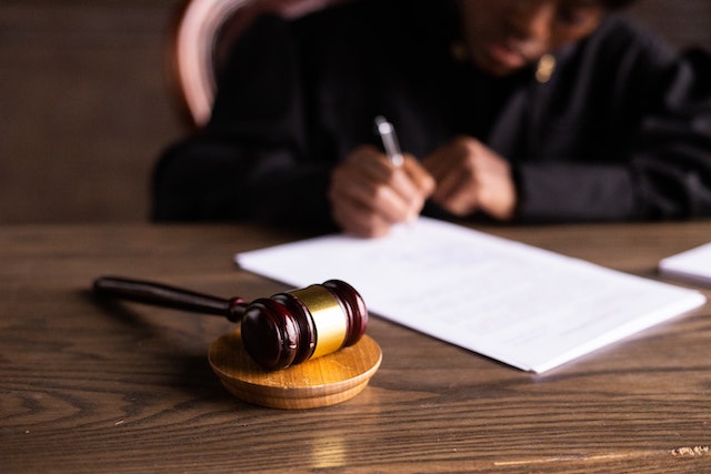 A wooden gavel rests on a desk in the foreground, while a person in a dark robe writes on papers in the background—a scene echoing courtroom decisions where Ignition Interlock Devices are often required in California.