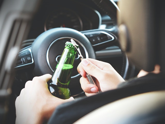 A person sitting in a car is opening a green glass bottle with a bottle opener while holding the steering wheel, highlighting potential road injury liability in California.