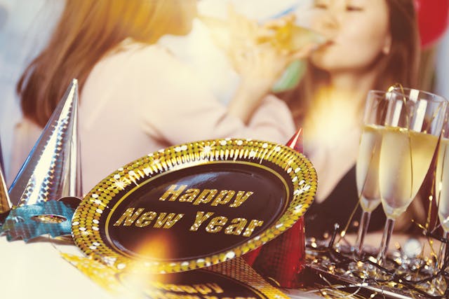 A close-up of a “Happy New Year” plate with party hats and champagne glasses, two people celebrating and drinking in the blurred background—reminding us of the serious California DUI penalties that can follow a festive night.