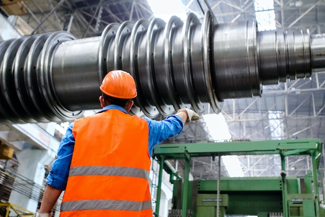 A worker in an orange safety vest and helmet inspects or gestures toward a large industrial machine component inside a factory, possibly following procedures related to a California Workers’ Compensation Appeal.