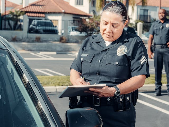 A police officer stands by a car writing on a notepad in a parking lot, preparing to file a police report after an accident in Sacramento, while another officer is visible in the background.