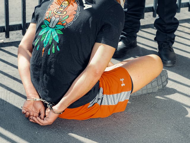 A person in an illustrated black T-shirt and orange shorts, possibly showing common concussion symptoms after a car crash, sits on the ground with hands cuffed behind their back near someone standing in black shoes.