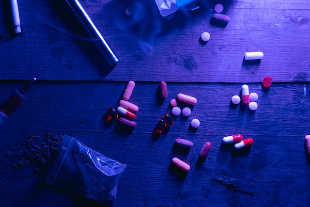 Assorted pills, capsules, a syringe, and a small plastic bag are scattered on a wooden surface under purple lighting, suggesting the presence of substances that could lead to a dispute over breathalyzer results in California.
