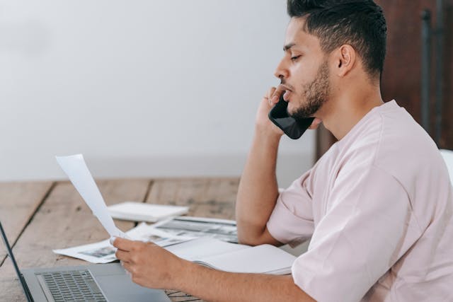 Man sitting at a desk holding a phone to his ear and looking at a sheet of paper, with a laptop and documents in front of him, possibly discussing how to dispute breathalyzer results for a California DUI case.