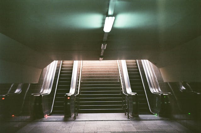 An underground passage in California with two escalators and a staircase leading up to daylight, lit by a single overhead fluorescent light—far from the bustling streets above filled with self-driving cars.