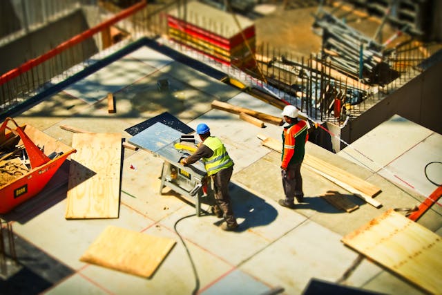 Two construction workers in safety gear operate a table saw and handle materials on a building site with scattered wooden boards and tools, illustrating potential scenarios for California premises liability claims.