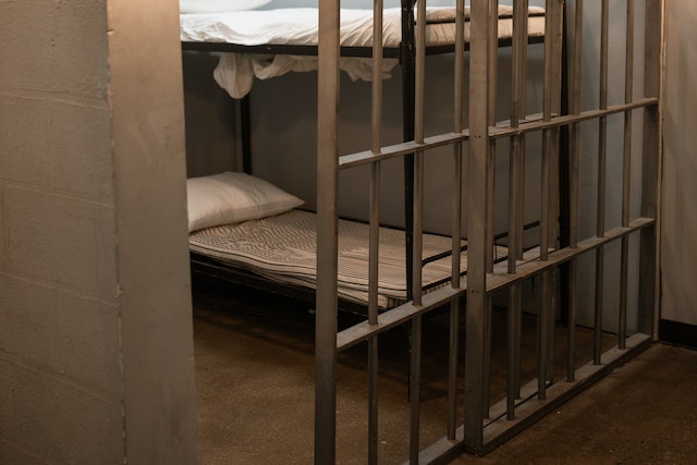 A small jail cell with metal bars contains a bunk bed with thin mattresses, a pillow, and white sheets on the lower and upper bunks, illustrating conditions that could raise California premises liability concerns.