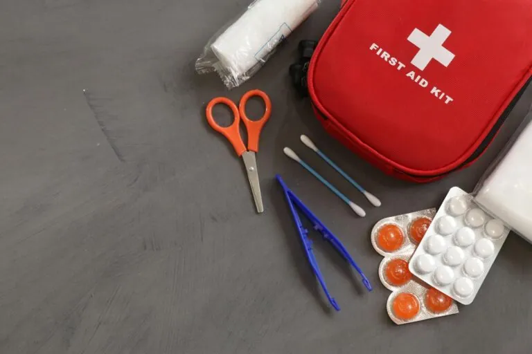 First aid kit with scissors, tweezers, cotton swabs, bandages, and blister packs of pills on a gray surface—essential supplies to minimize the impact of injuries or while awaiting an injury claim.