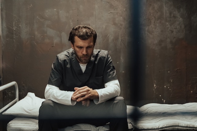 A man in prison uniform sits on a bed in a jail cell, looking down with his hands clasped, appearing deep in thought about what to do if falsely accused of domestic violence in California.