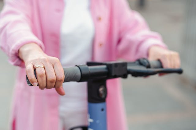 A person wearing a pink jacket grips the handlebars of an electric scooter in California, with a gold ring visible on one hand.