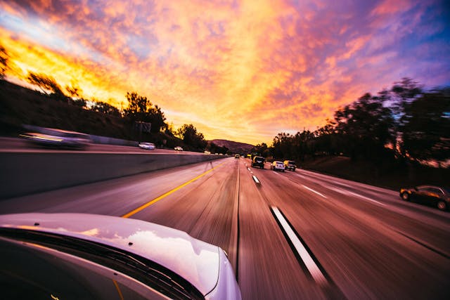 A self-driving car speeds down a multi-lane California highway at sunset, with blurred motion and vibrant orange and purple clouds glowing in the sky.