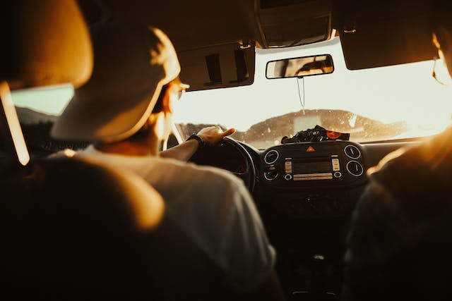 Person wearing a backwards cap drives a car at sunset, with sunlight streaming in through the windshield and car dashboard visible—a reminder to recognize common symptoms of concussion after any car crash.