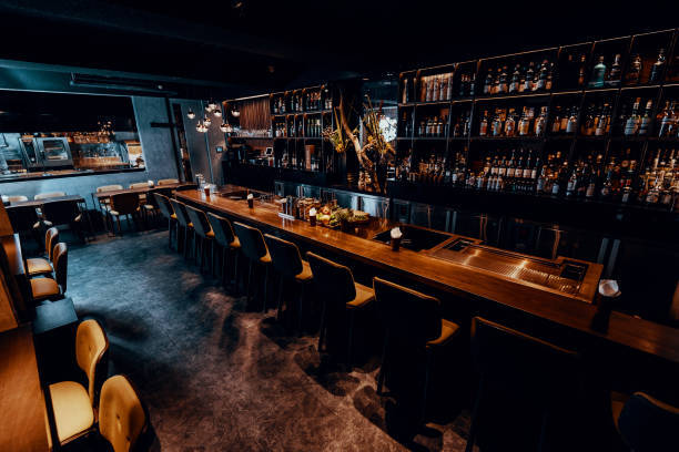 Modern bar interior with a long wooden counter, high bar stools, and shelves stocked with bottles of liquor; dim lighting creates a cozy atmosphere but could pose a risk of poor lighting injury if not careful.