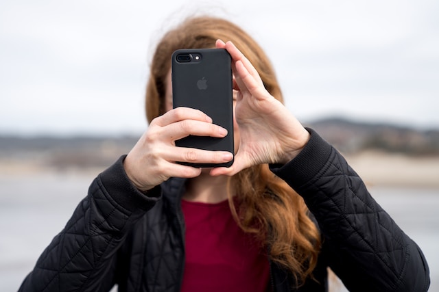 A person with long hair wearing a black jacket holds a smartphone up to take a photo outdoors, obscuring their face—perhaps documenting dangerous road conditions California is known for.